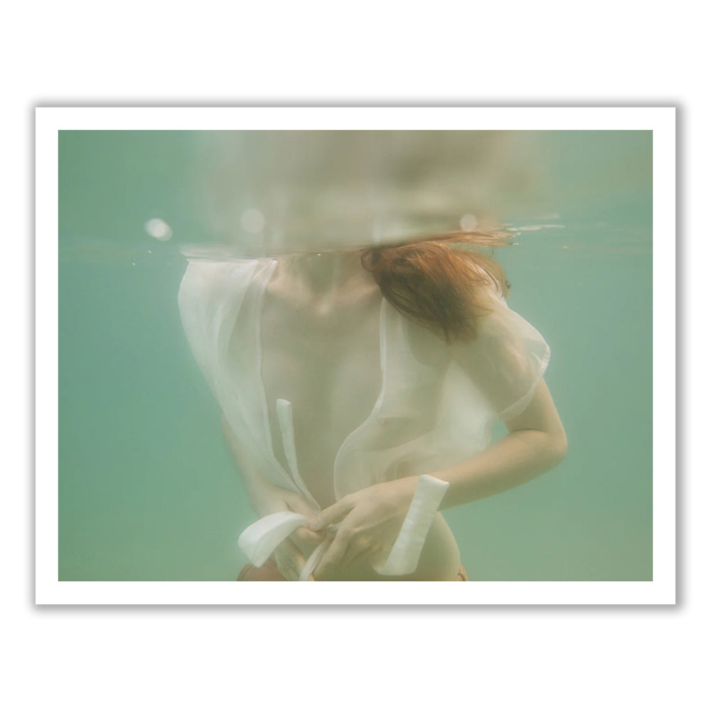 Underwater photo of a person with long hair wearing a partially transparent, flowing white garment. The upper part of their body and face are submerged, creating a soft, dreamy effect with the water's surface blurring the image. This fine art piece, titled "Softness #1" by Francesca Owen, is printed with archival pigments on quality paper for durability.