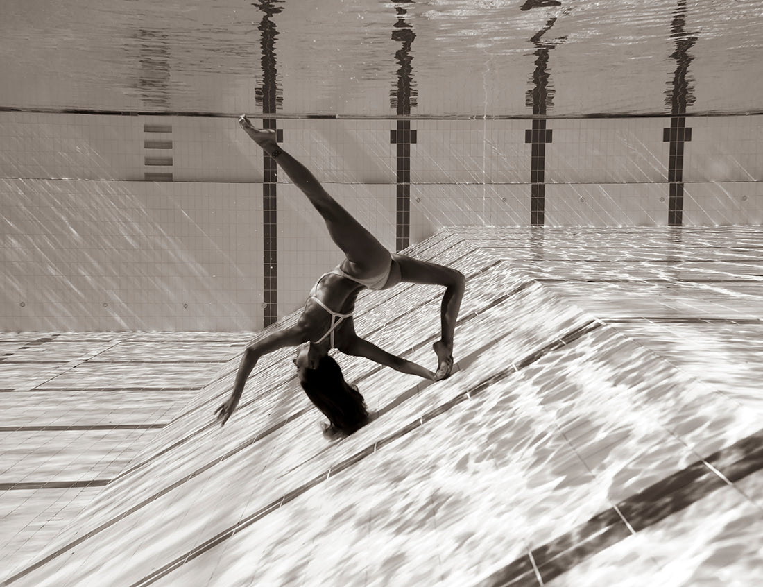 In Francesca Owen's "Flying Solo 04," a person underwater in a pool is gracefully posed in an elegant, arched backbend position on the sloped pool floor. The black-and-white image, printed on fine art paper with archival pigments, captures the rippling water and the reflection on the pool's tiled surface.