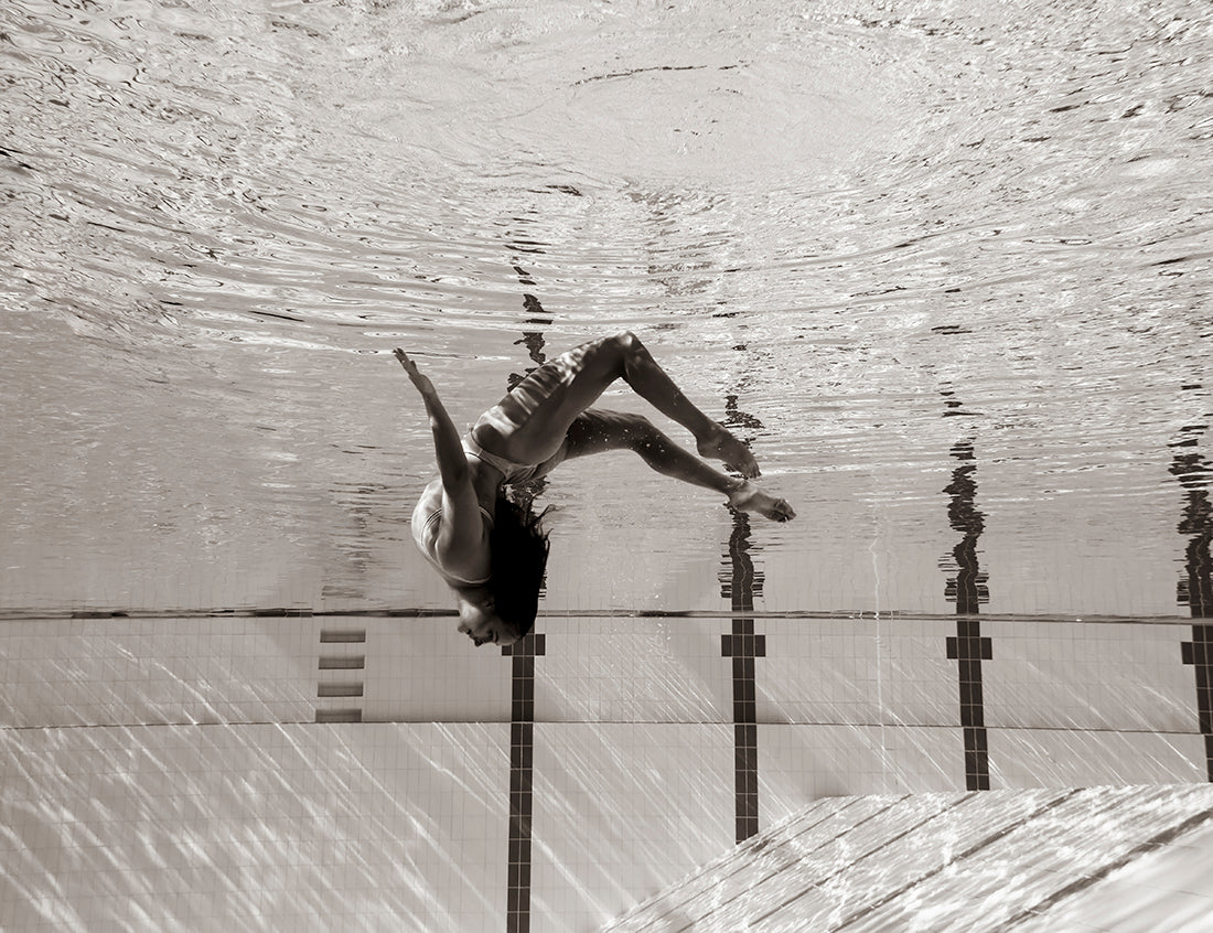 In the stunning image "Flying Solo 07" by Francesca Owen, a person underwater in a pool strikes a graceful, upside-down pose with bent legs and extended arms, creating dynamic ripples. The background reveals the pool's ladder and tiled wall, with reflections dancing on the water's surface. This captivating piece is available in custom sizes.
