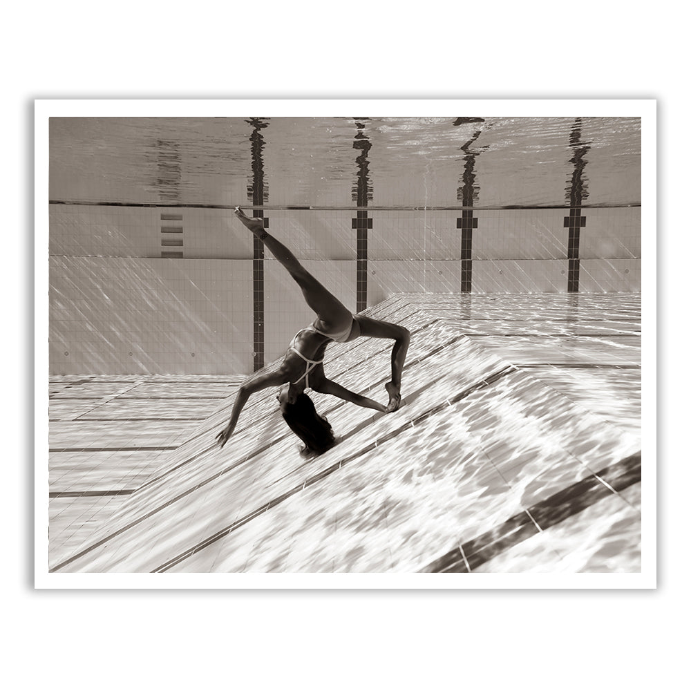 Flying Solo 04 by Francesca Owen features a sepia-toned photograph of a person in a one-piece swimsuit performing an elegant underwater pose in a swimming pool. The image, printed on fine art paper with archival pigments, captures the subject with one arm extended downward and one leg lifted while touching the pool floor, creating mesmerizing reflections on the water surface above. Each print comes with a certificate of authenticity.