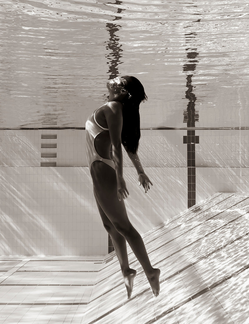 Product Description: Francesca Owen's "Flying Solo 05" captures a stunning black and white underwater photo of a woman in a swimsuit, gracefully floating towards the water's surface with her back arched and arms extended outwards. The pool tiles and surface reflections create mesmerizing patterns. Printed on fine art paper using archival pigments, it comes with a certificate of authenticity.