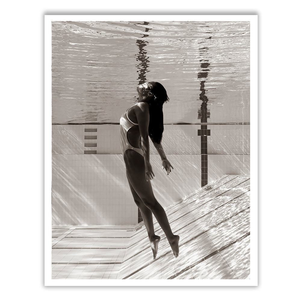 Flying Solo 05 by Francesca Owen captures a black-and-white underwater photograph of a woman in a one-piece swimsuit. She is submerged in a swimming pool, facing upward and appearing to float gracefully near the pool's edge where light patterns are visible on the water surface and pool tiles. Printed on fine art paper with archival pigments, this piece includes a certificate of authenticity.