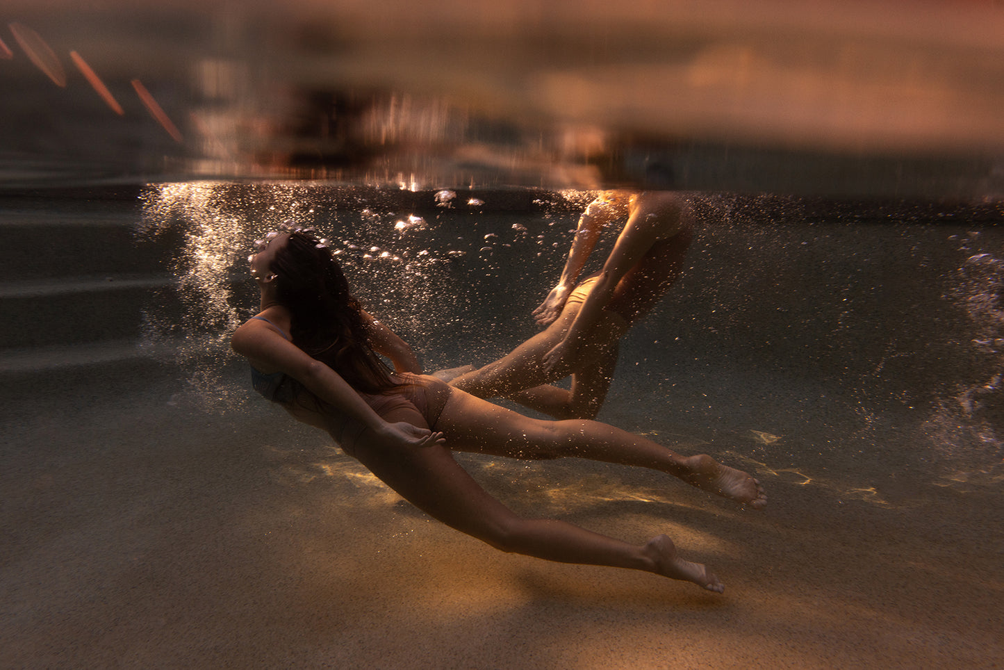 Two people swim underwater in a pool, surrounded by bubbles and warm light with stairs on the left. Sunset #1 by Francesca Owen is available as a fine art print with certificate of authenticity.
