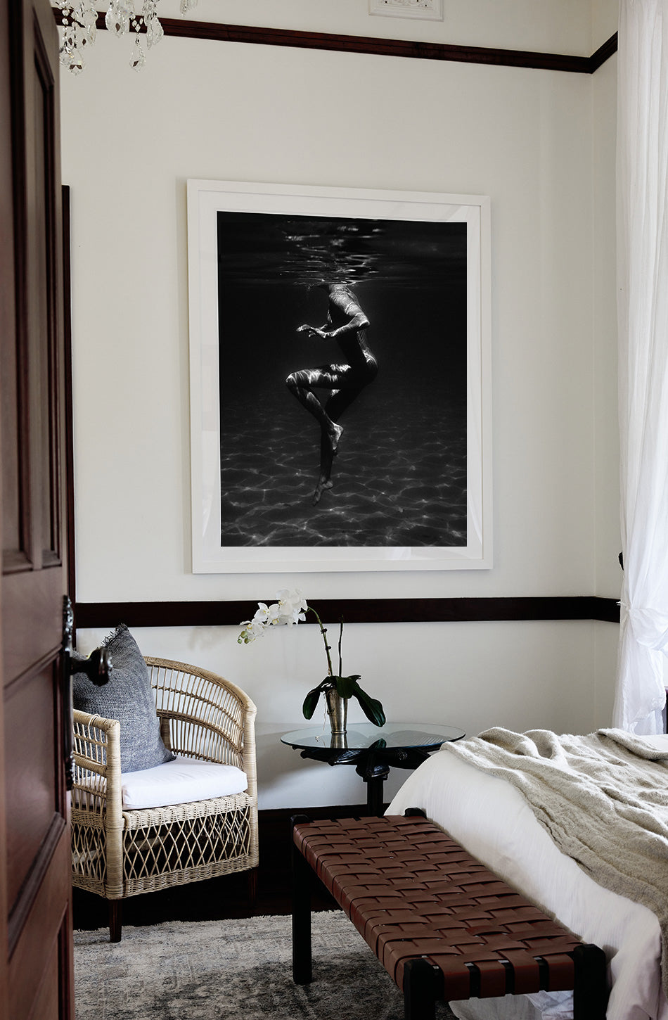 A cozy bedroom with a wicker chair, a glass side table displaying an orchid, and a bench situated at the foot of the bed. Hanging above the chair is "Still Waters #1," an elegant large black and white photograph on fine art paper by Francesca Owen, featuring an underwater scene with a swimmer. Soft light filters through white curtains.