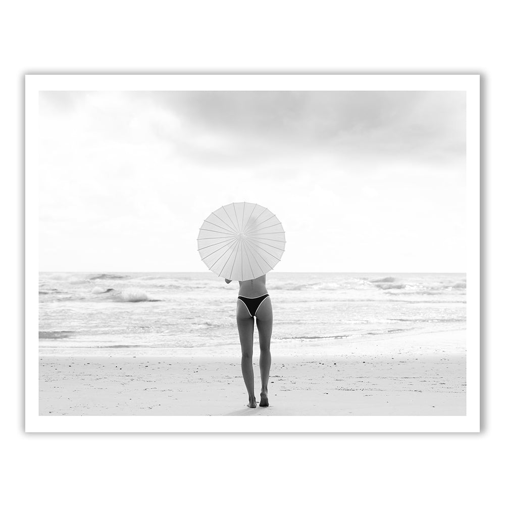 The Beauty Within Part 2 #3, a captivating black and white photo by Francesca Owen, captures a person in a bikini standing on the beach and facing the ocean while holding a parasol. The overcast sky and gently crashing waves create an ambiance of tranquility and solitude. This serene scene is printed with archival pigments on fine art paper.