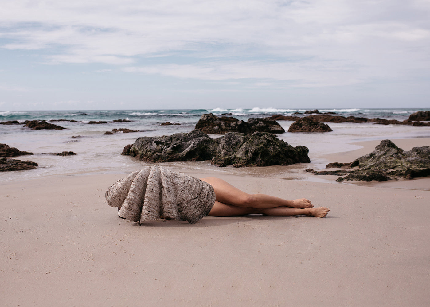 A person lies on a sandy beach, partially wrapped in a large seashell, with the ocean and rocks visible in the background under a cloudy sky. This captivating scene, titled "The Beauty Within" by Francesca Owen, is captured using archival pigments and available on fine art paper with worldwide shipping.