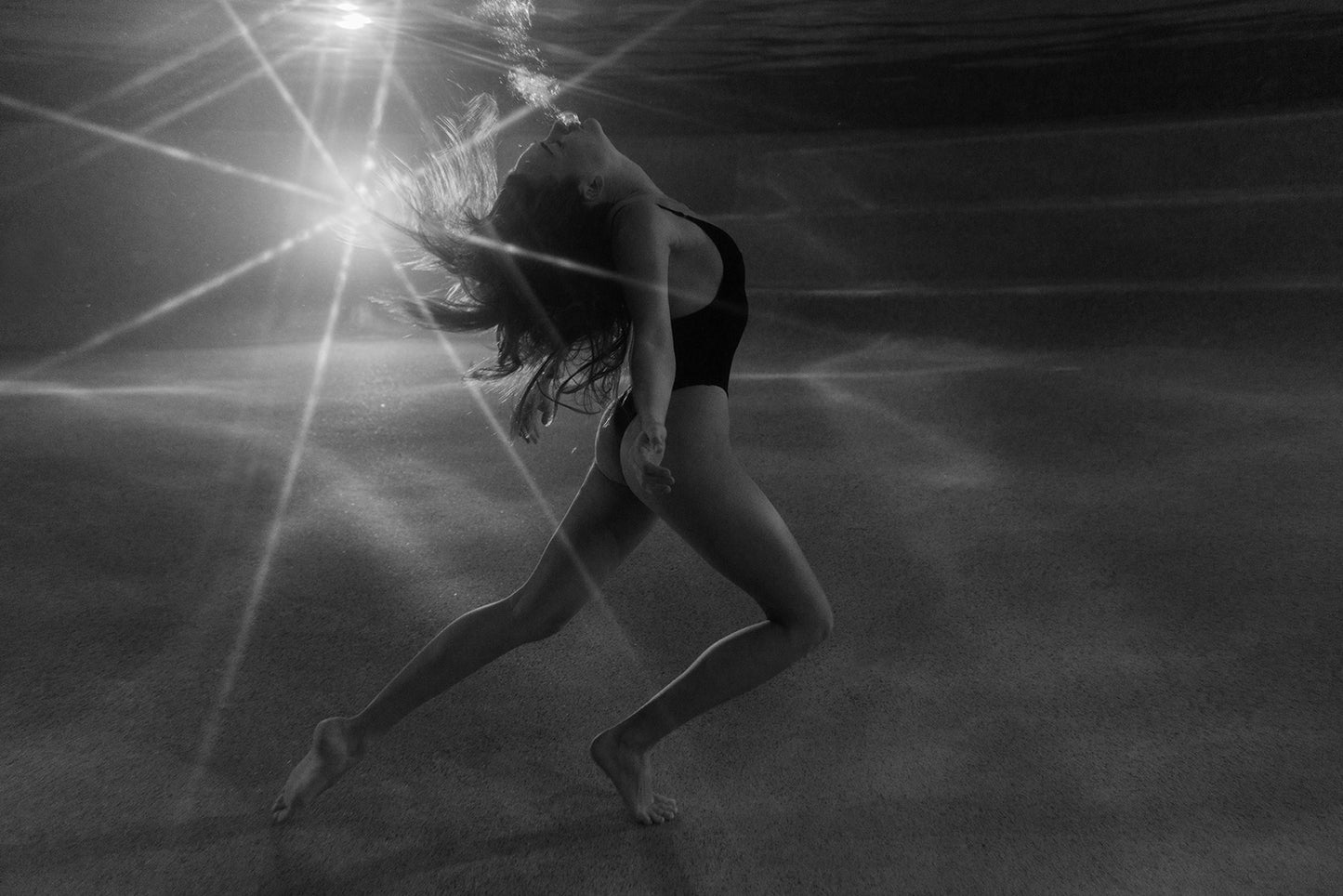 A woman in a swimsuit poses gracefully underwater with her hair flowing amid rays of light. "The Bower #2" by Francesca Owen is a dreamy black and white fine art print and includes a certificate of authenticity.