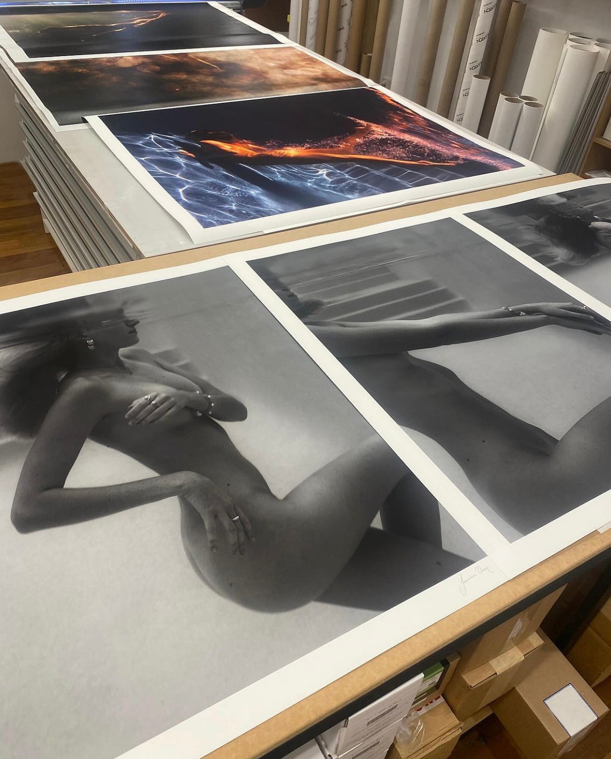On a table, large unframed prints are displayed with two black-and-white prints from Francesca Owen's collection titled "Unveiled 02," featuring a nude figure in an abstract pose on fine art paper. In the background, other prints showcase vibrant, abstract designs. Rolls of paper are neatly stacked on the sides of the table, accompanied by a certificate of authenticity.