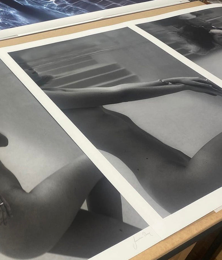 Francesca Owen's 'Unveiled 03' fine art prints, featuring black and white images of a nude figure, are elegantly displayed on a table. The photos, aligned side by side, showcase different angles and parts of the body in an artistic composition that focuses on form and light. Printed with archival pigments for longevity, these artworks come with a certificate of authenticity.