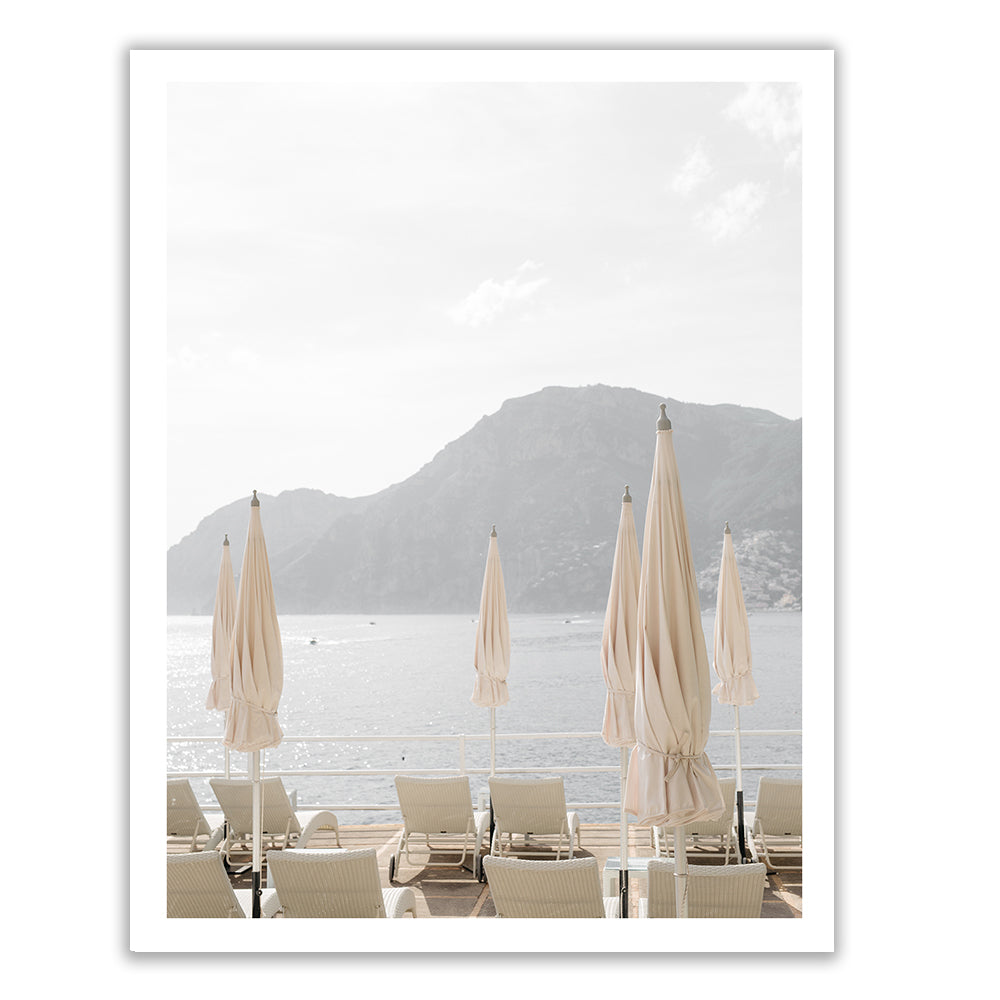 A serene seaside view featuring Francesca Owen's hand-signed "Umbrellas #2" with beige umbrellas and lounge chairs facing a calm, sunlit body of water. In the background, a mountainous landscape rises against the soft sky. The scene, printed with archival pigments on fine art paper, conveys a peaceful, relaxing atmosphere.