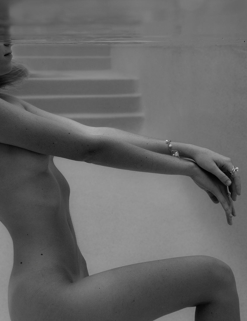 Francesca Owen's "Unveiled 03" captures a serene, black and white image of a person submerged in water up to their chest. The individual, either sitting or kneeling underwater, extends their arms gracefully in front of them. Bubbles and reflections enhance the tranquility of the scene, while a bracelet and rings adorn their hands. This captivating piece is available as fine art prints with archival pigments.