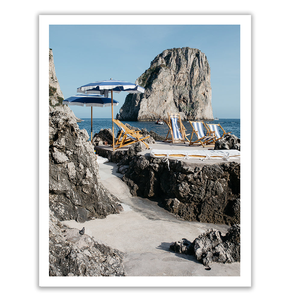 The artwork "Waiting For You #1" by Francesca Owen showcases a coastal scene with rocky outcrops, a sandy area adorned with blue and white striped deck chairs and matching umbrellas. In the background, a large rocky island rises from the calm blue sea under a clear sky. This elegant scene is captured on fine art paper using archival pigments and is available for worldwide shipping.