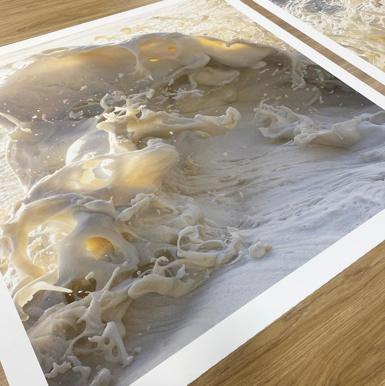 A close-up of the limited edition "Wild Seas Part I, V I" print by Francesca Owen showcases frothy ocean waves on a sandy shore. The foam and water create swirling patterns and textures, yielding an abstract, almost sculptural appearance. This archival pigment print is elegantly displayed on a wooden surface.