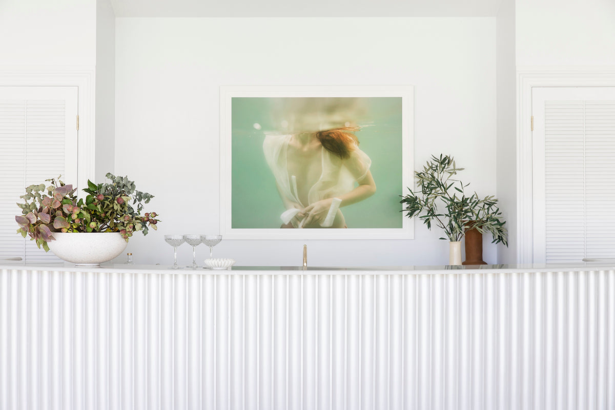 A minimalist white room with a fluted countertop. On the countertop are a large bowl of greenery and a smaller plant in a brown pot. Above the counter, there's "Softness #1," a framed photograph by Francesca Owen printed on fine art paper featuring a person underwater wearing a white shirt, complete with an archival pigments certificate of authenticity.