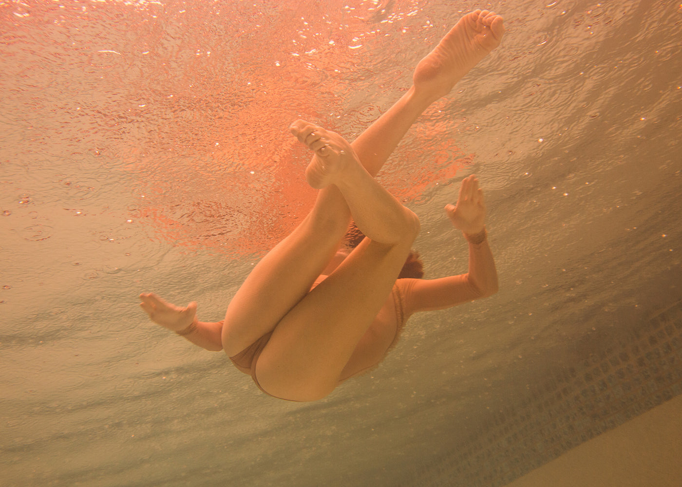 A person submerged in a swimming pool strikes a dynamic pose with their legs and arms visible. The rippling water surface above them is illuminated by an orange-tinted light, revealing the pool tiles in the background. This image, titled "Another World," is part of the Limited Edition collection by Francesca Owen and is available with worldwide shipping.