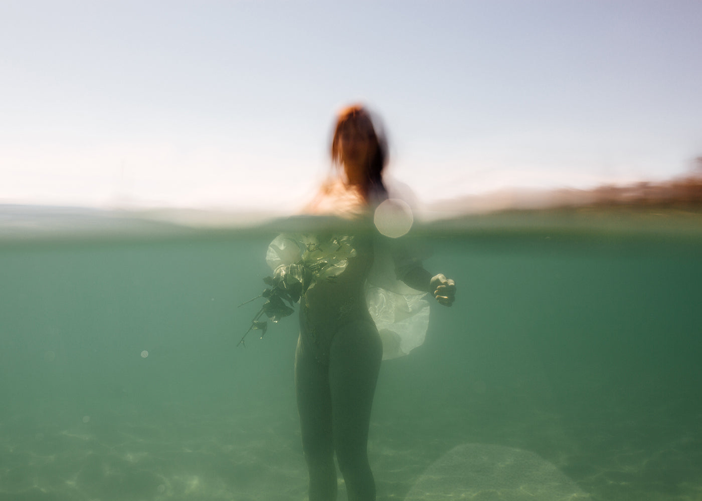 A dreamlike and serene artwork titled "The Language of Flowers #3" by Francesca Owen depicts a blurred image of a person standing partially submerged in clear, green-tinted water, holding a bouquet. The top half showcases a clear sky and distant shoreline. This fine art piece is printed with archival pigments on fine art paper and is hand-signed for authenticity.