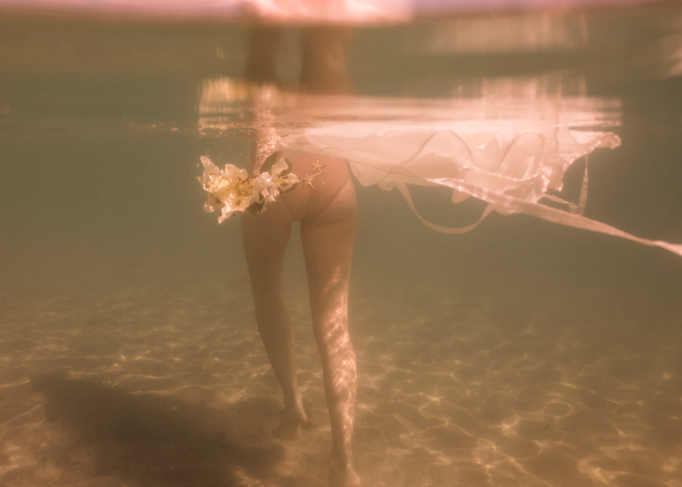 Underwater view of a person in a white dress walking through clear water with a sandy bottom. The dress floats around as they hold a small bouquet of white flowers. Sunlight filters down, casting golden reflections, making it perfect for capturing as "The Language of Flowers #2" by Francesca Owen in fine art prints, all accompanied by a certificate of authenticity.
