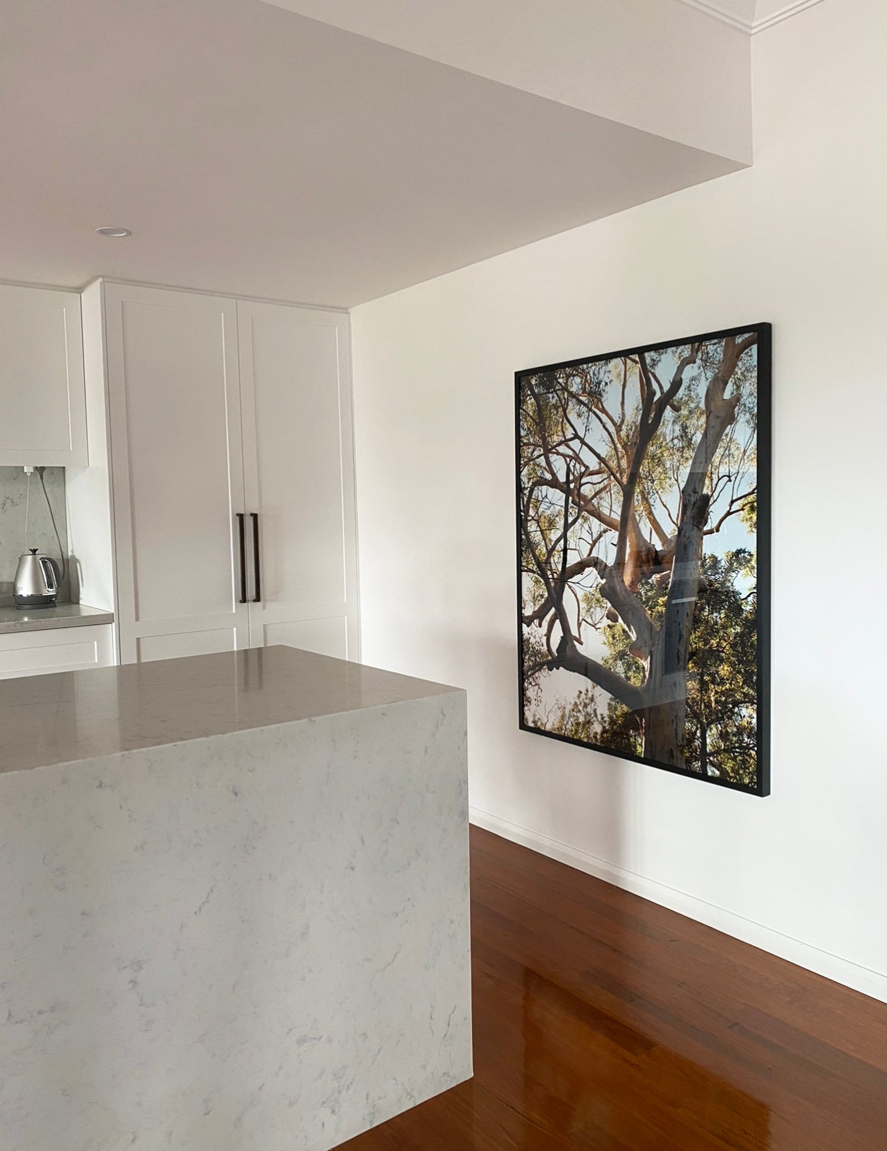 A modern kitchen with white cabinetry features a large marble island countertop. A stainless steel kettle sits on the counter in the background. On the adjacent white wall hangs "Australia #1" by Francesca Owen, a fine art print certified with archival pigments depicting a tree against a bright sky. The floor is polished wood.