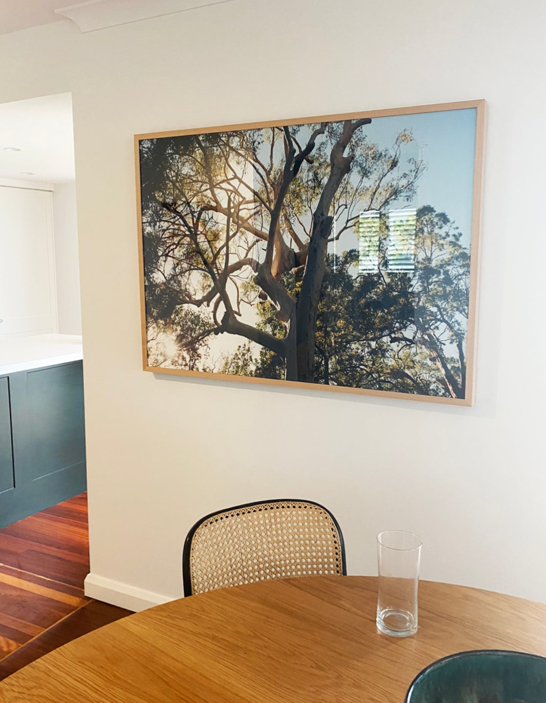 A dining area featuring a wooden table, a glass vase, and a chair with a woven backrest. Mounted on the white wall above the table is "Australia #1" by Francesca Owen, a large framed photograph of a sunlit tree printed on fine art paper and accompanied by a certificate of authenticity. The adjacent kitchen area is partially visible.