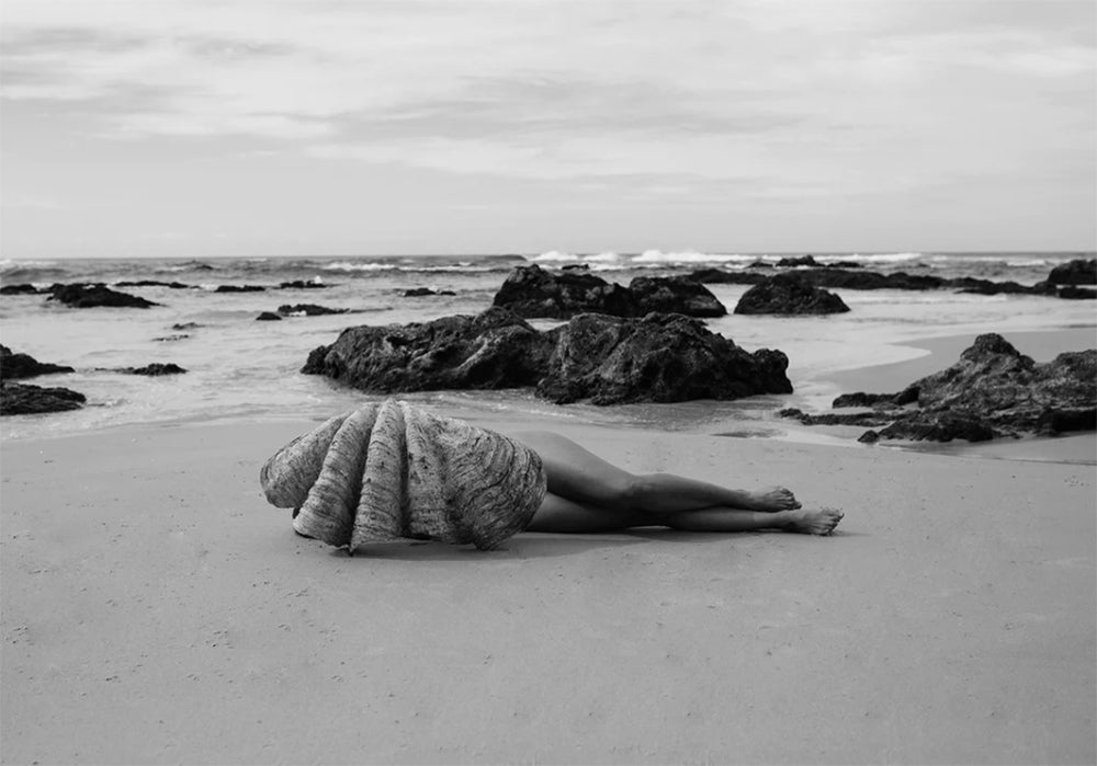 **Product Description:**
"The Beauty Within B&W" is an exquisite fine art photograph by Francesca Owen. This black and white masterpiece captures a beach scene with rocks scattered near the shoreline. A person lies on the sand, mostly covered by a large shell, facing away from the camera as waves gently lap at the shore under an overcast sky. This stunning piece is available with worldwide shipping.