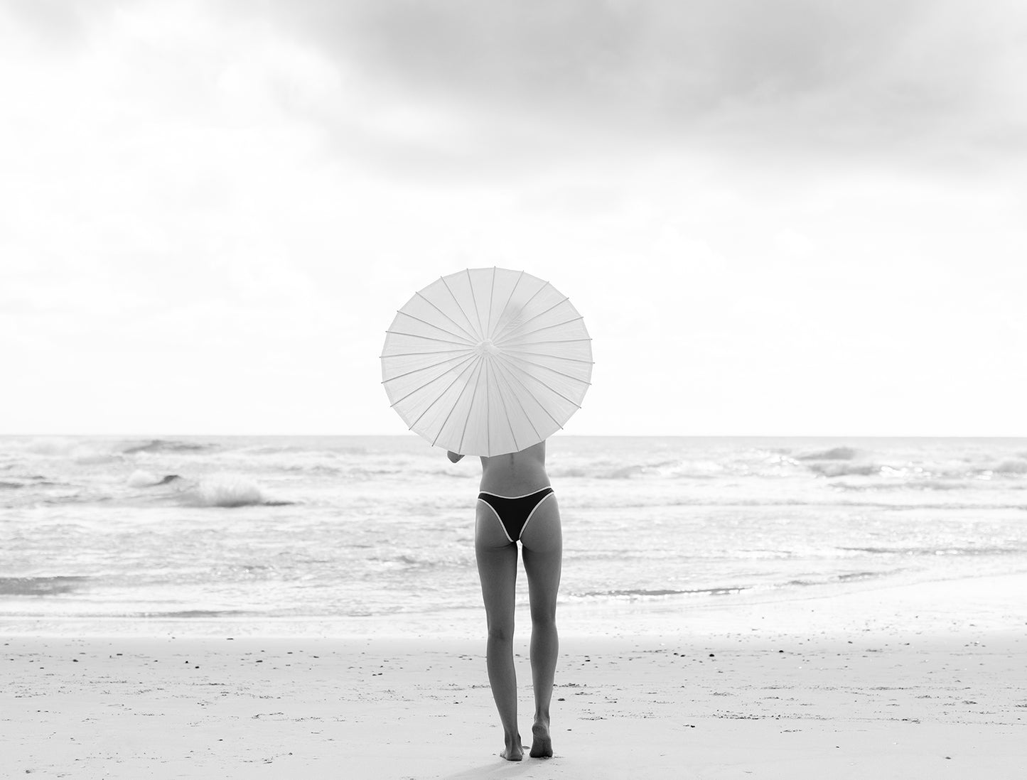 A person in a bikini stands on the beach facing the ocean, holding a white parasol and appearing to gaze at the distant waves. The sky is cloudy, and the scene unfolds in black and white tones, akin to "The Beauty Within Part 2 #3" by Francesca Owen, expertly captured using archival pigments on fine art paper.