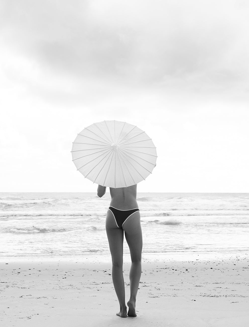A person stands on a beach facing the ocean, holding a white parasol above their head. They are wearing black swimwear and the scene is captured in black and white, perfect for printing on fine art paper. Waves gently crash on the shore in the background, evoking serenity. This captivating moment is beautifully rendered by Francesca Owen in The Beauty Within Part 2 #2.
