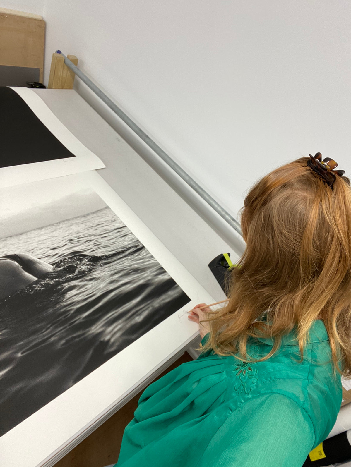 A woman with long red hair, wearing a green top, is standing at a table signing "Bottoms Up," a large black-and-white photograph of a wave by Francesca Owen. The setting appears to be an art studio with drawers underneath the table and a wooden shelf in the background, where fine art prints tagged with certificates of authenticity are displayed.