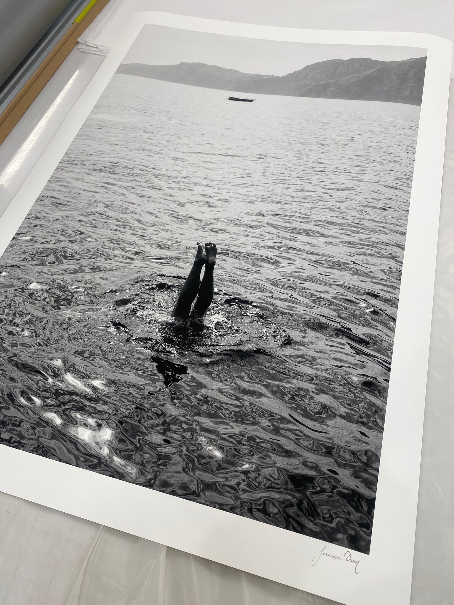 Capri Tippy Toes" by Francesca Owen captures a striking black and white image of a person performing a handstand in the water, with only their legs and feet visible above the surface. In the background, distant mountains and a solitary boat float on calm waters. This piece is available in custom sizes with archival pigments and includes a certificate of authenticity.