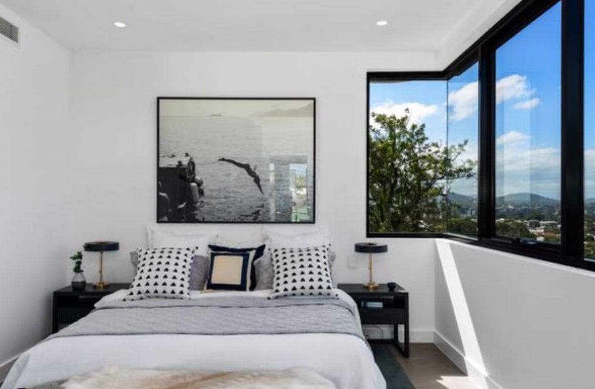 A modern bedroom features a bed adorned with monochrome pillows and a grey throw. Above the bed hangs "Dive In," a large framed photo from Francesca Owen, printed on fine art paper with archival pigments, depicting a person standing near a diving board. A corner window with black frames offers clear views of trees, hills, and a blue sky with clouds.