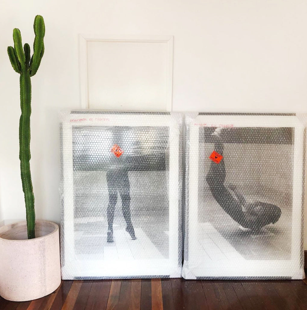 Two large, framed Tippy Toes photos wrapped in bubble wrap lean against a white wall beside a tall cactus in a white pot. Each photo, printed on fine art paper with archival pigments by Francesca Owen, has an orange tag attached. The framed photos appear to depict people in various poses. The floor is made of dark wood.
