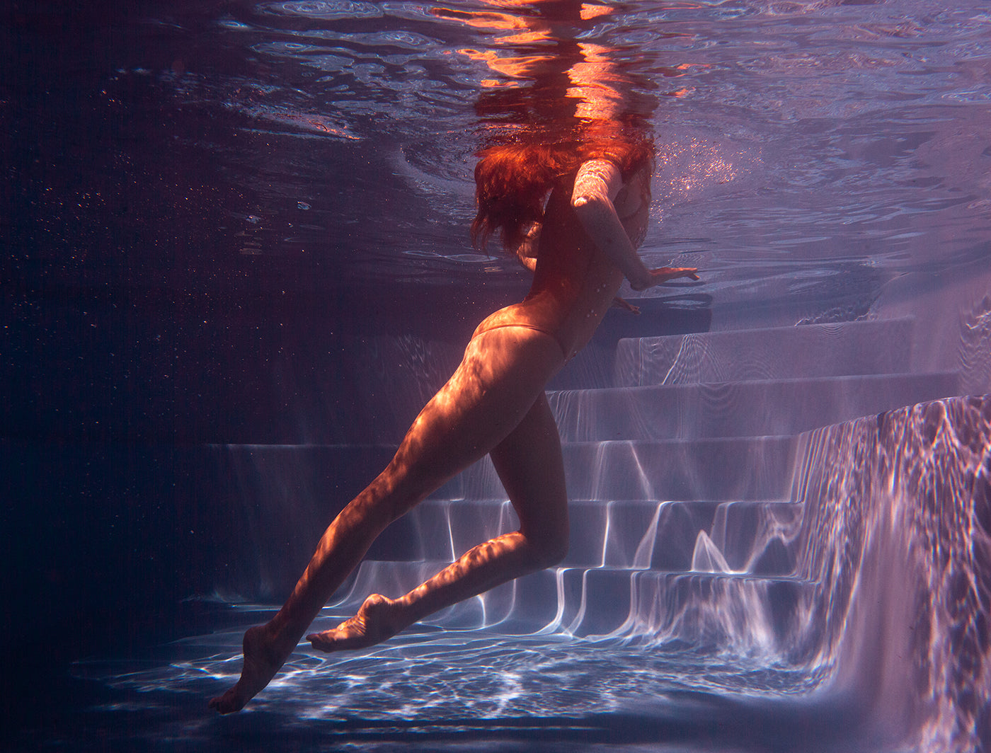 A person with long red hair is swimming underwater near a set of steps. The light creates rippling patterns on their skin and the surrounding water, giving the scene an ethereal, dreamlike quality, reminiscent of fine art prints captured with Francesca Owen's Glisten 04 archival pigments.
