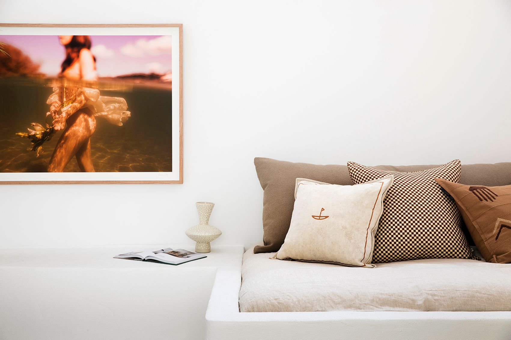A minimalist room features a white couch with beige and brown accent pillows. A framed, artistic photograph titled "The Language of Flowers #1" by Francesca Owen, printed with archival pigments on fine art paper, hangs on the white wall above the couch. A white table beside the couch holds an open book and a small, textured vase.