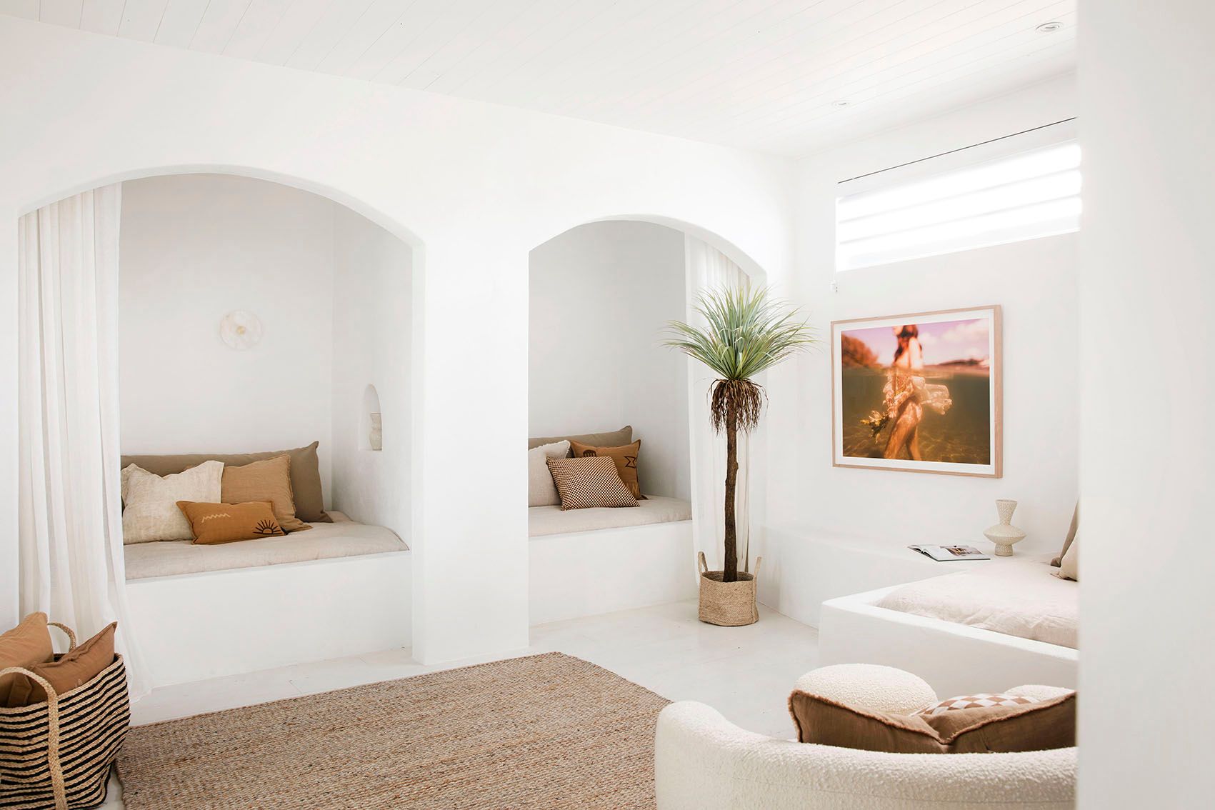 A minimalist white living room features built-in alcove seating with neutral-toned cushions and throw pillows. A tall indoor plant and a woven rug add warmth, while "The Language of Flowers #1" by Francesca Owen, framed on fine art paper, and soft natural light from a window enhance the cozy atmosphere.