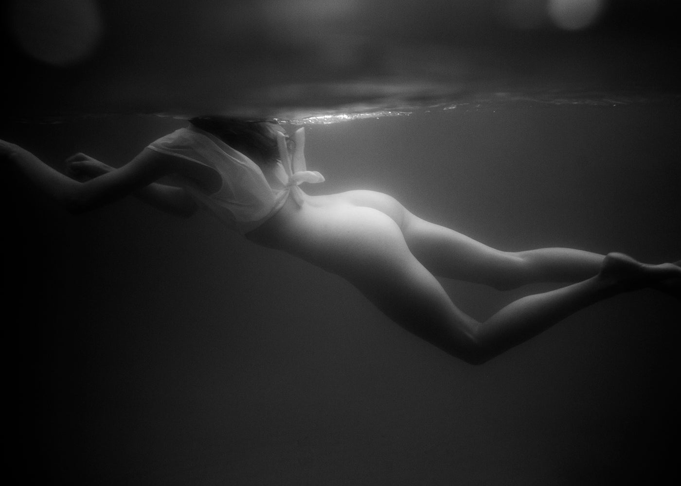 Gliding #1 by Francesca Owen captures a side view of a person swimming underwater, floating horizontally in a light-colored top with no visible bottom attire. The water's surface is subtly rippled, creating delicate light patterns. This black and white image is available in custom sizes and printed with archival pigments. Each piece comes with a certificate of authenticity.