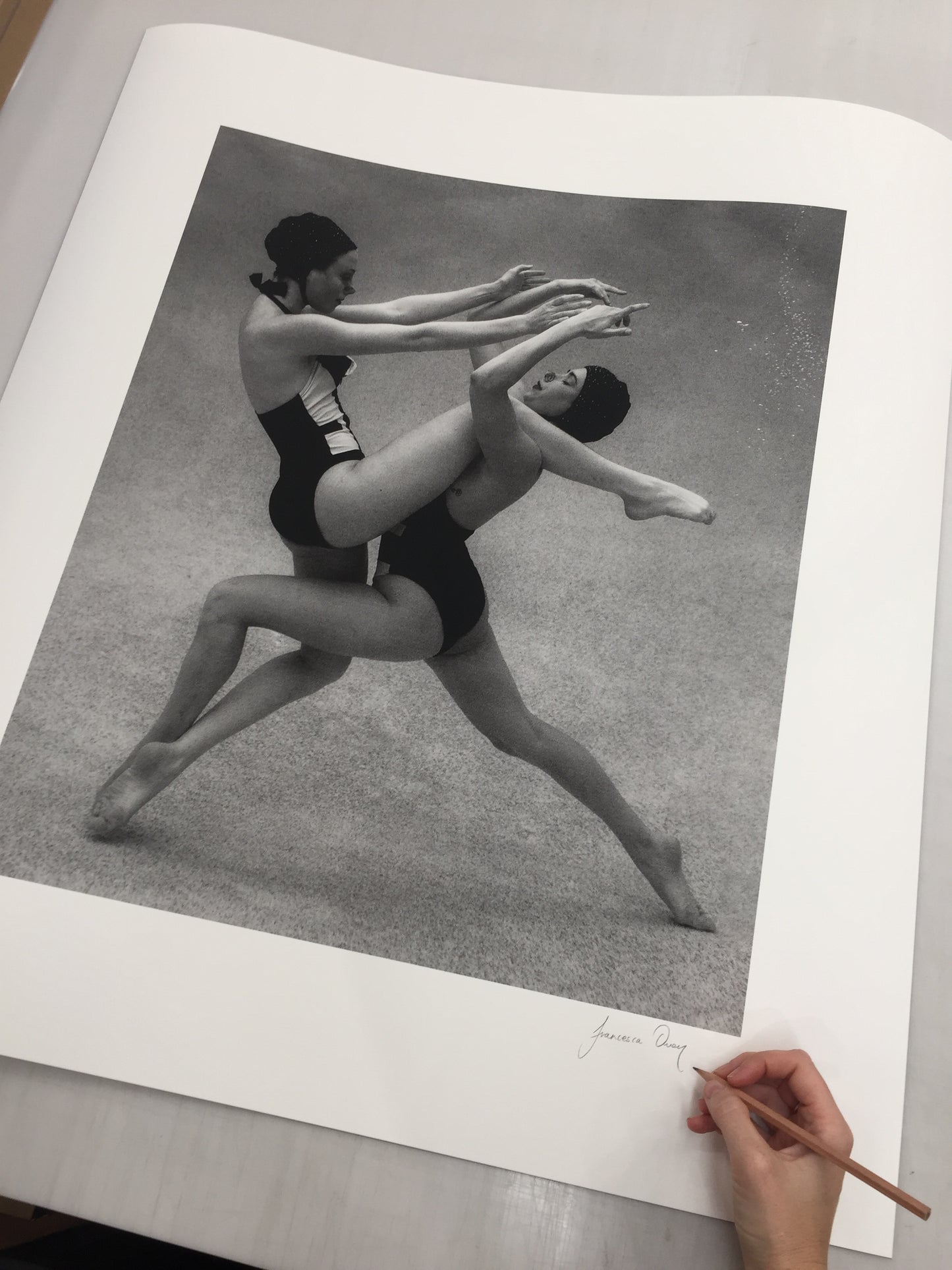A person is holding a pencil and about to sign "Palm Springs Duo #1," a large black-and-white photograph by Francesca Owen, printed on fine art paper. The photograph features two synchronized swimmers in mid-air performing a routine, wearing swim caps and swimsuits. Complete with a certificate of authenticity, the photo is laid out on a table.