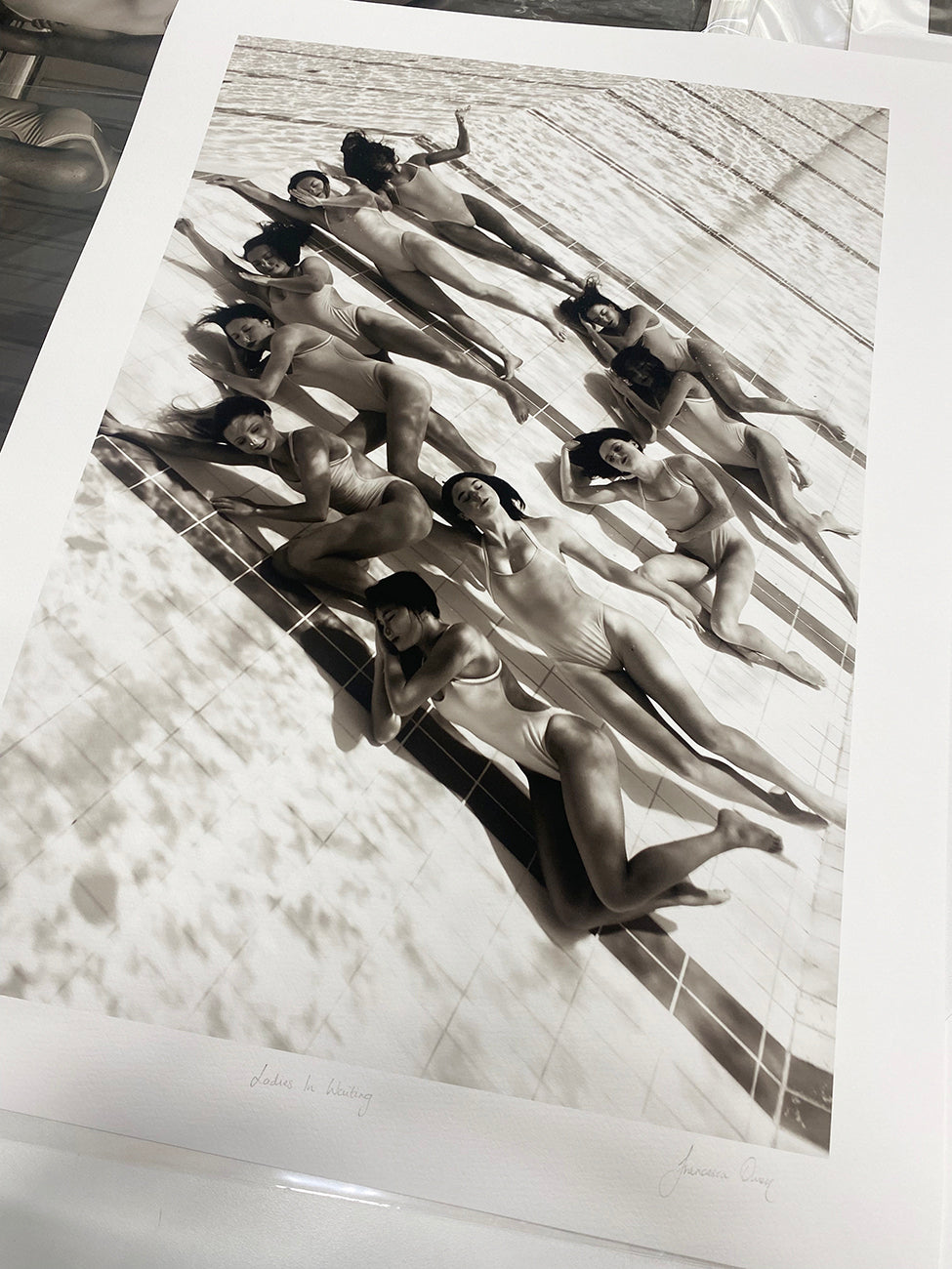 Francesca Owen's 'Ladies in Waiting #2' is a captivating black and white photograph that beautifully captures several women lying side by side, sunbathing by the edge of a pool. Positioned in a staggered, overlapping manner, they create an artistic pattern reminiscent of synchronized swimming. This fine art print is available as a large limited edition display on a flat surface.