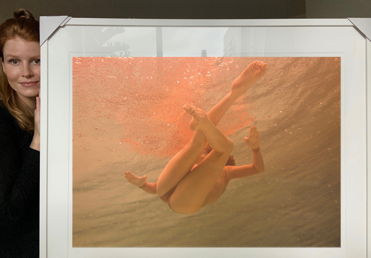 A person with long red hair stands to the side, holding a framed, hand-signed photograph. The image depicts a submerged individual underwater in a bent, floating position, surrounded by warm, orange-tinted water. This piece titled "Another World" by Francesca Owen is part of a Limited Edition collection available for worldwide shipping.
