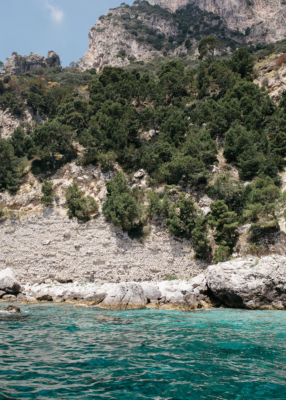 Introducing "Aqua Waters #2" by Francesca Owen: A beautiful depiction of a rocky shoreline with clear turquoise water, leading up to a steep, tree-covered hillside. The sky is clear and blue, and the rugged landscape showcases layers of rock and greenery, creating a natural yet dramatic scene. This stunning piece is available in custom sizes and framing options.