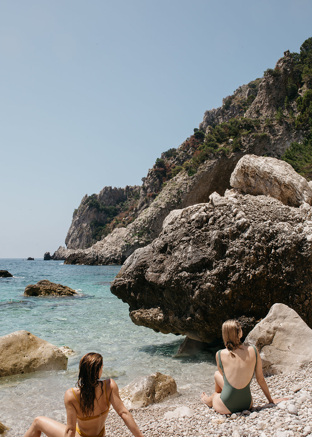 Two people sit on a rocky beach, facing a serene turquoise sea. Rugged cliffs covered with greenery rise up in the background under a clear sky. The person on the left wears a yellow bikini, and the person on the right wears a green one-piece swimsuit. The scene is reminiscent of "Capri Mermaids" by Francesca Owen, as if captured in fine art prints with archival pigments.