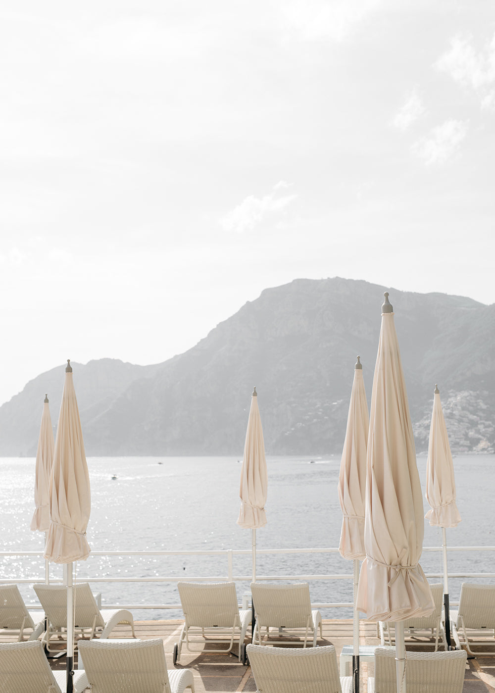 Experience the serene and inviting mood of Francesca Owen's artwork, "Umbrellas #2." This fine art piece, printed on premium quality paper using archival pigments, captures a tranquil beach scene. Beige closed umbrellas and empty lounge chairs face calm waters, with hazy mountains rising in the background against a light, overcast sky.