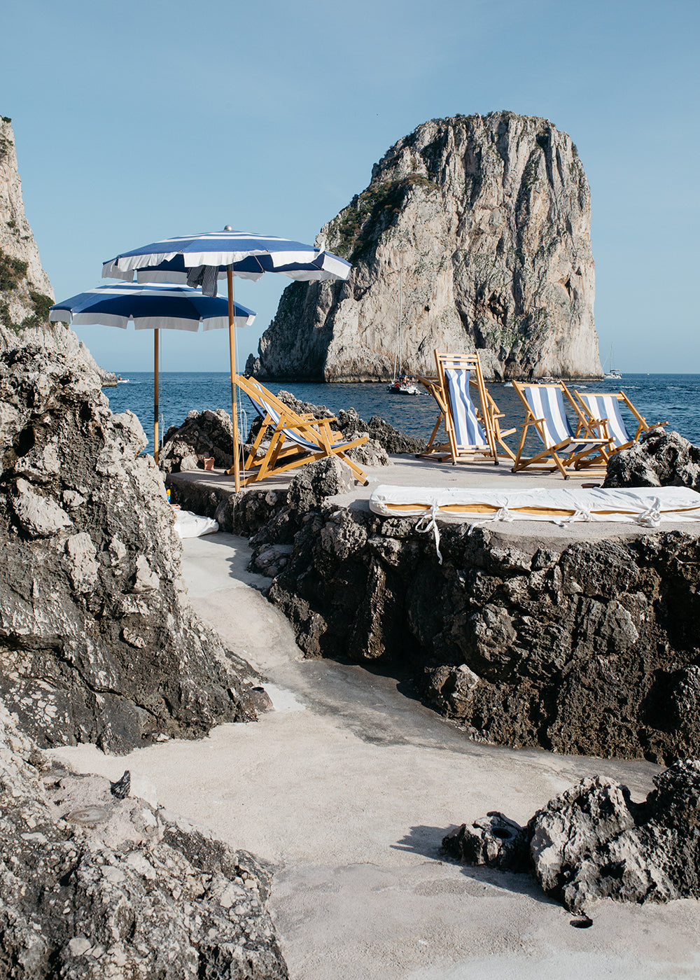 The artwork "Waiting For You #1" by Francesca Owen showcases a coastal scene with rocky outcrops, a sandy area adorned with blue and white striped deck chairs and matching umbrellas. In the background, a large rocky island rises from the calm blue sea under a clear sky. This elegant scene is captured on fine art paper using archival pigments and is available for worldwide shipping.