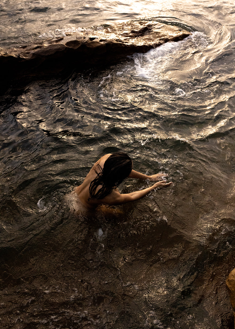 A person with long dark hair is immersed in swirling, shallow water near a rock formation. The scene, bathed in the warm light of sunset, reflects beautifully on the water's surface. The individual interacts with the water, creating ripples akin to waves of archival pigments on a canvas titled *Sunrise Swim #9* by Francesca Owen.