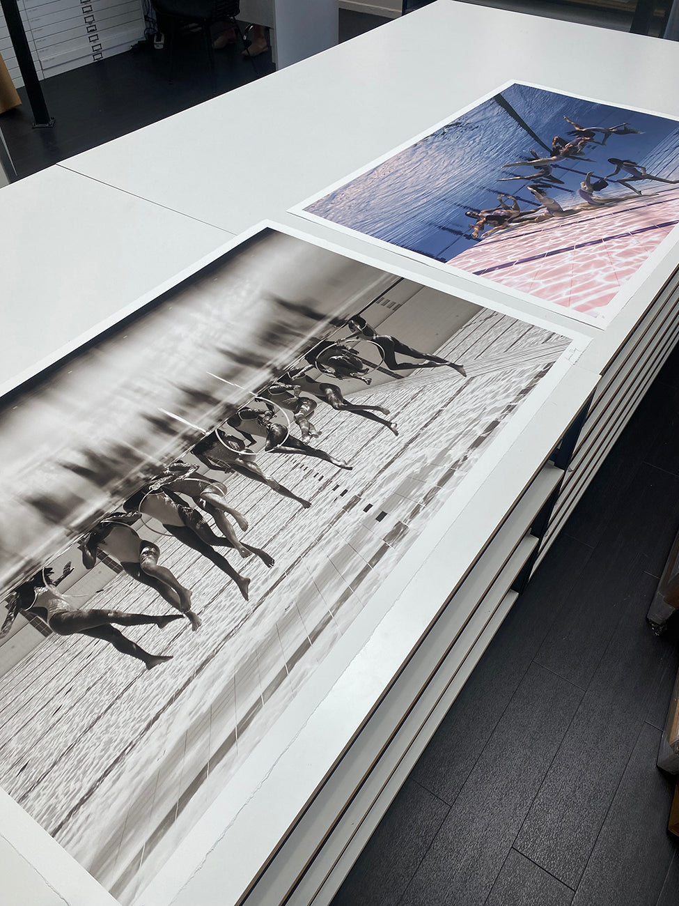 Two large photographs displayed on a table showcase the stunning work of fine art photographer Francesca Owen. The left photo, printed on Giclee fine art paper, is a black-and-white image of people swimming underwater. The right, titled "Ladies in Waiting #3," is a limited edition color photograph that captures swimmers racing in individual lanes from above.