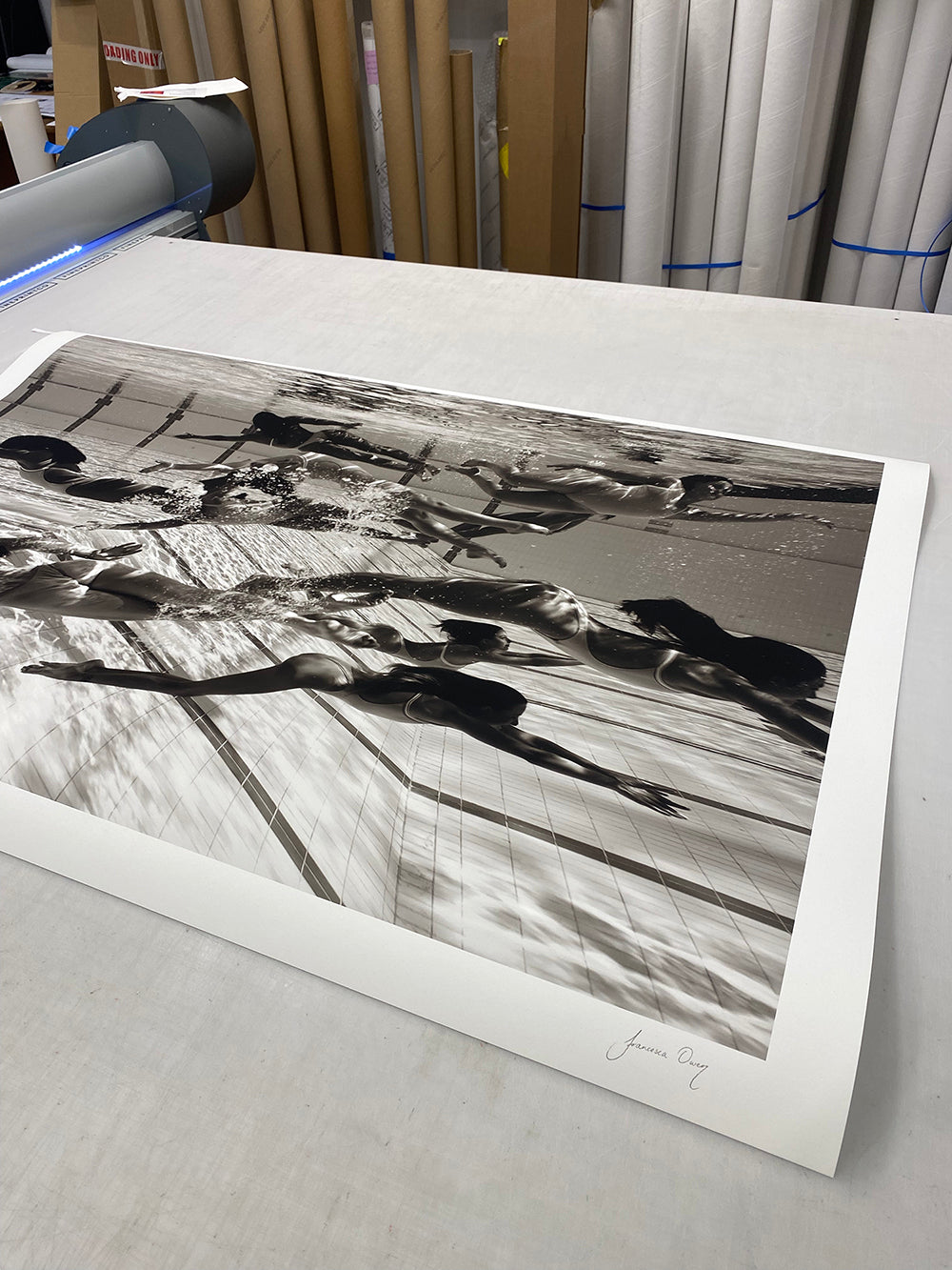 A limited edition black and white photograph titled "Ladies in Waiting #4" by Francesca Owen, featuring swimmers underwater executing various strokes, is laid out flat on a table for printing. The print is signed by the fine art photographer in the lower right corner. Rolls of giclee fine art paper prints and various printer components are visible in the background.