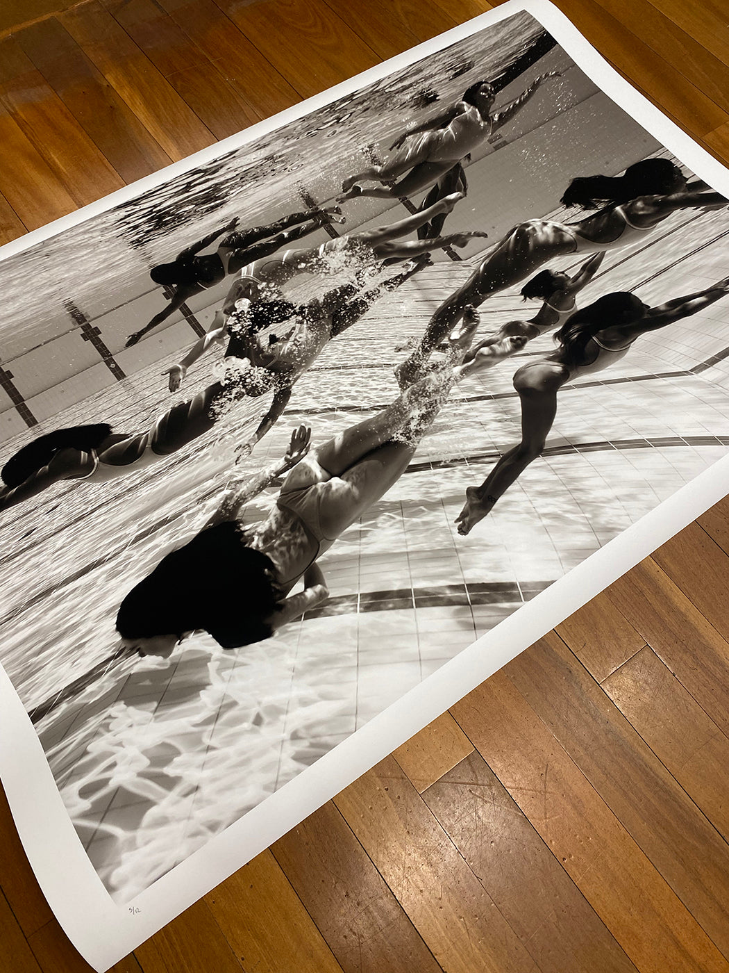 A black and white photograph titled "Ladies in Waiting #4" by Francesca Owen lies on a wooden floor, depicting underwater swimmers in a pool. Captured on Giclee fine art paper by this fine art photographer, the image showcases dynamic motion and the play of light on the water's surface, with legs and arms in various positions of movement.