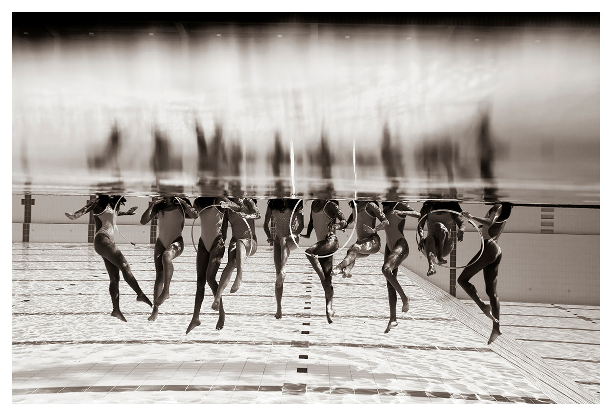 A synchronized swimming team performs upside-down in a swimming pool, showcasing their legs and torsos perfectly reflected on the water's surface. Aligned in a row and holding circular hoops, they create a visually stunning symmetrical effect. Captured by fine art photographer Francesca Owen, this mesmerizing scene is available as a limited edition print titled "Ladies in Waiting #3" on giclee fine art paper.