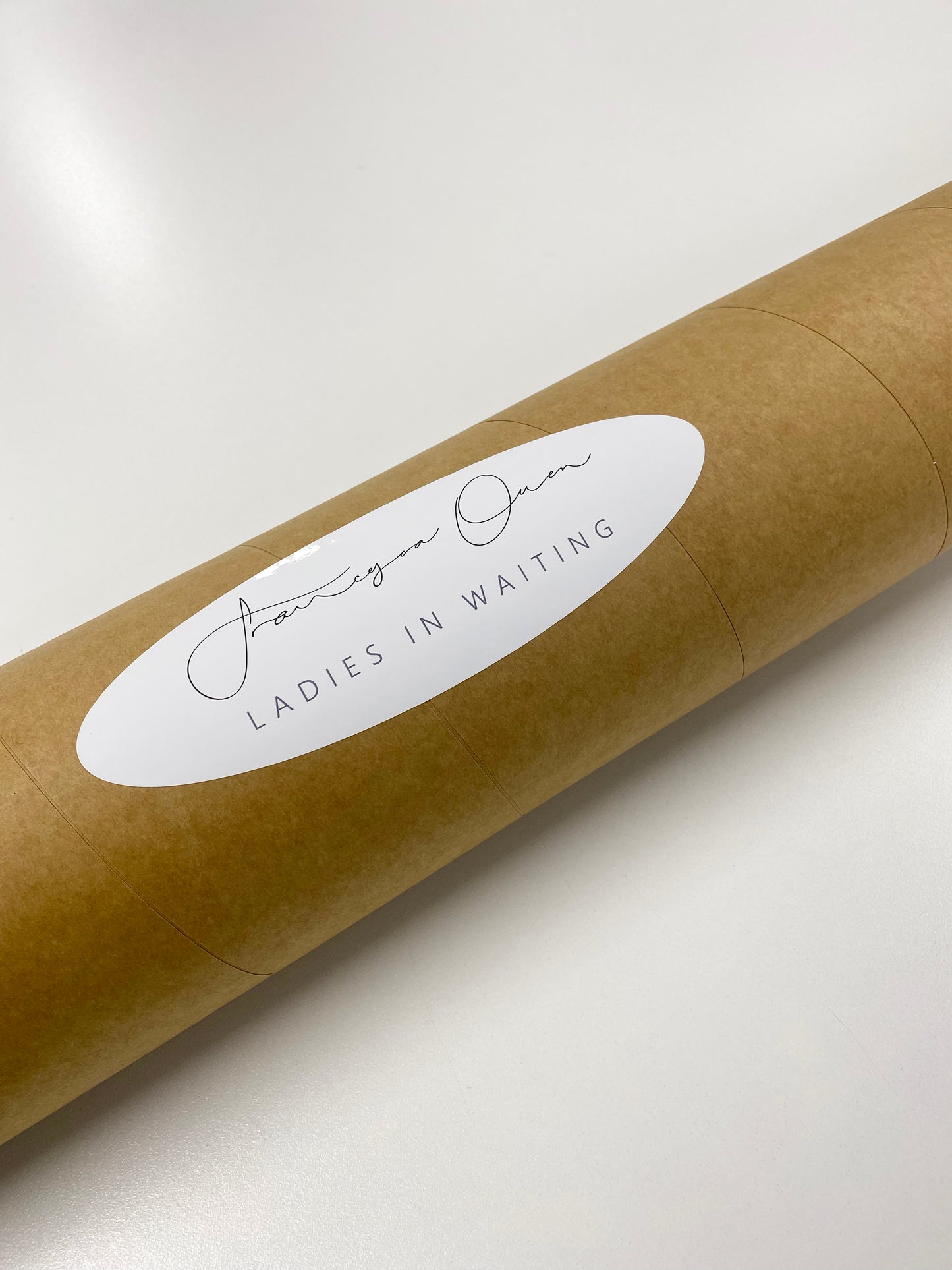 A brown cylindrical tube is displayed on a white surface, featuring a white oval label adorned with cursive text that reads "Ladies in Waiting #2" beneath the signature of Francesca Owen. This indicates it is part of a Limited Edition series. The tube appears to be designed as a mailing or storage container, possibly intended for housing works on giclee fine art paper.