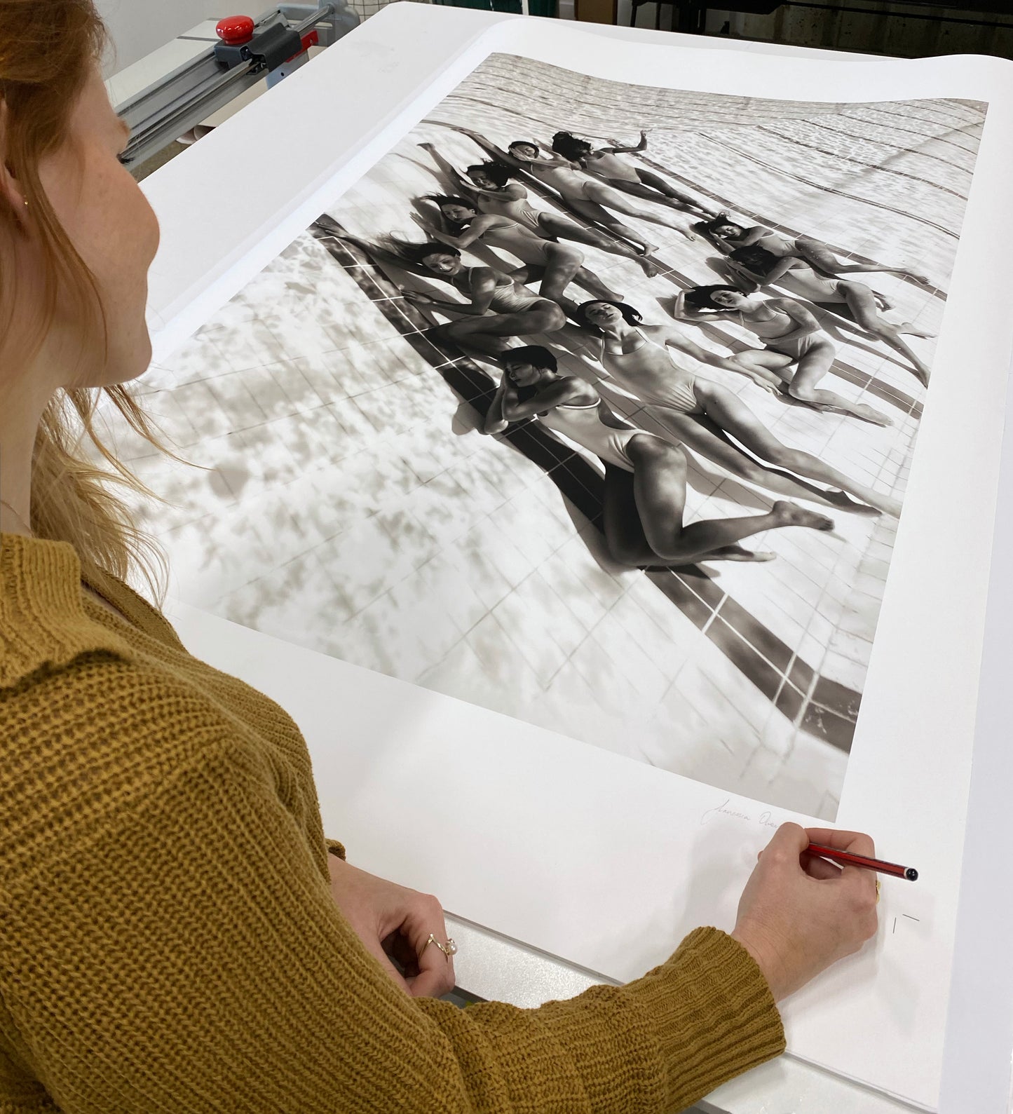 A person in a mustard-yellow sweater signs a large black-and-white Fine Art Print titled "Ladies in Waiting #2" by Francesca Owen, which features a group of individuals in swimsuits posing underwater. The person, partially visible from the side, holds a red pencil while finalizing this Limited Edition piece.