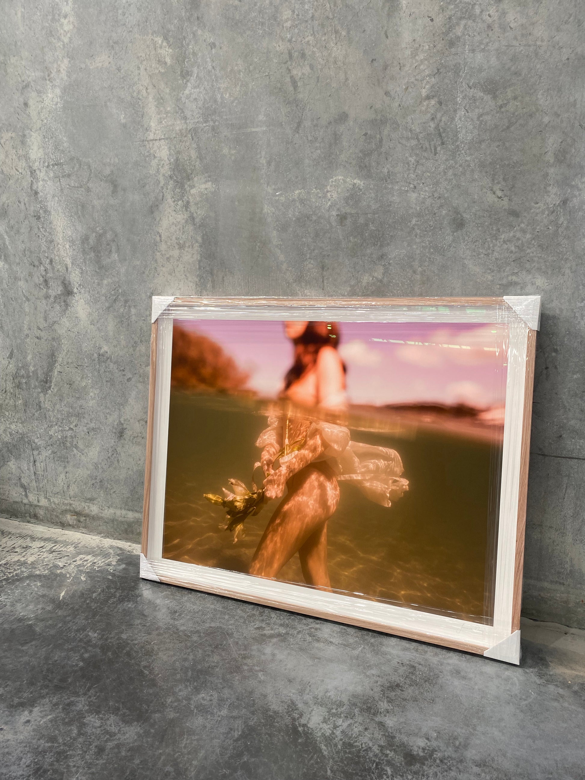 A framed photograph titled "The Language of Flowers #1" by Francesca Owen, set against a gray concrete wall, depicts a woman underwater holding flowers. Wrapped in protective plastic with cardboard corners, it suggests newness or transit. Printed on fine art paper with archival pigments, this serene and ethereal piece includes a certificate of authenticity.