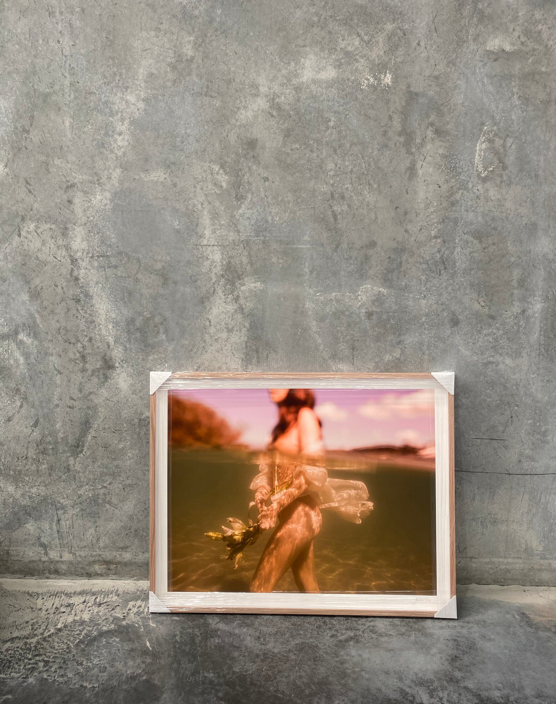 The Language of Flowers #1 by Francesca Owen is a framed photograph featuring a warm-toned, blurry image of a person partially submerged in water, printed on fine art paper with archival pigments. The indistinct background of trees and sky adds depth to the piece, while the frame rests against a gray textured concrete wall.
